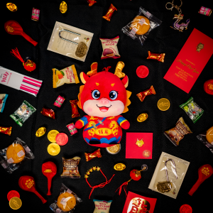 Assorted Dragon Box goodies shown scattered around a red and gold plush dragon. Goodies include special red lunar new year balloons, chocolate coins, red envelopes, lucky candy, and other sweets.