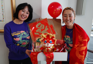 CCAI co-Founder and CEO Lily Nie and The Park Executive Director Amy Zhong are wearing the exclusive JCCS Year of the Dragon longsleeve shirts and holding an open Dragon Box, displaying some special items inside. Visible items include a red and gold plush dragon, red new year balloons, cashmere dragon scarf, CCAI water bottle, blessing character, and uninflated mylar balloon.